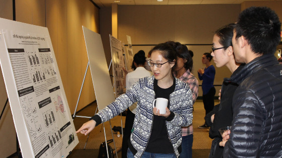 A woman holding a coffee cup speaks, pointing at a poster, while two people look on; behind them are more posters and people.