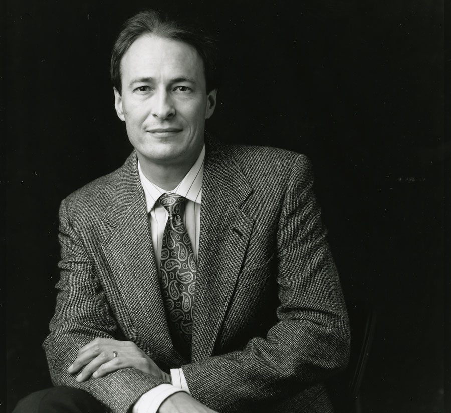 A crisply focused black and white photo of a man sitting in a suit and tie, smiling, leaning slightly to the left