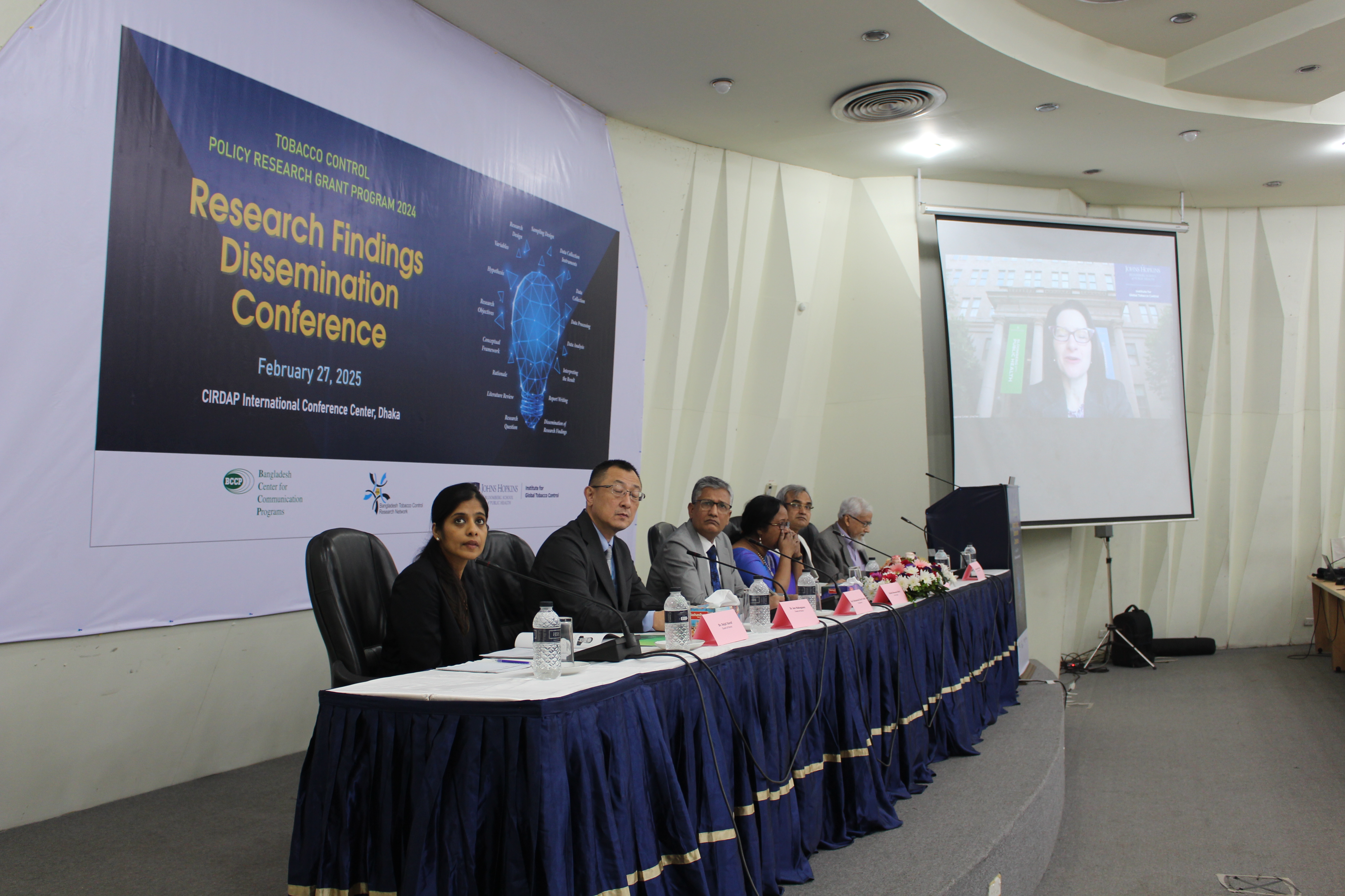 A group of people on the dais listen to a woman speaking via video to a conference.