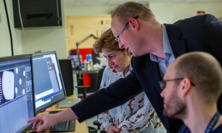 Three people face a desk with a pair of computer monitors on the left. Associate Professor Scott Bailey leans forward, reaching to point at the computer screen. Behind him to the left, Elizabeth Walder stands, looking down at section of the screen that Bailey is pointing at. On the right, in front of Bailey, postdoctoral fellow Evan Worden sits, head turned toward the screen.