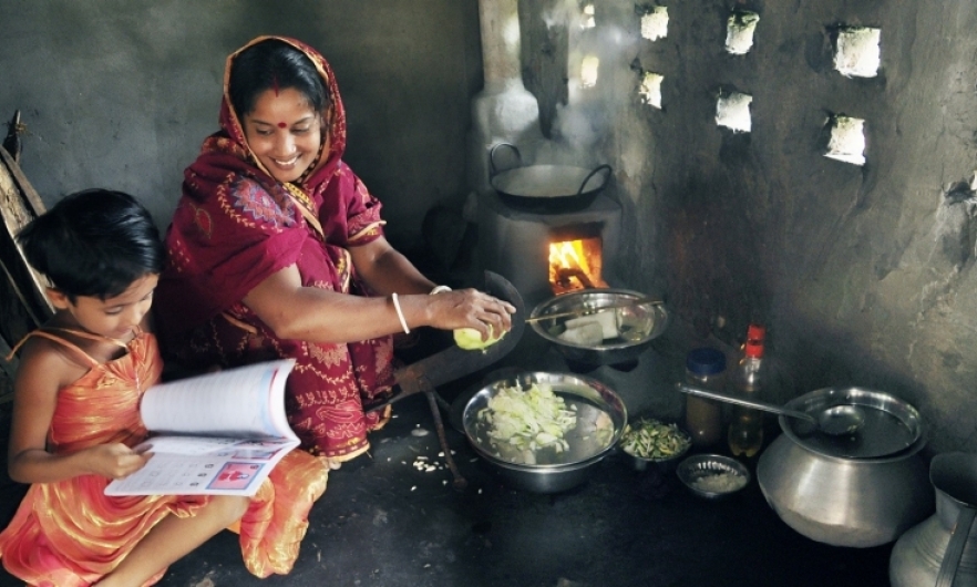 Woman in Dhaka uses a cookstove while a child next to her reads