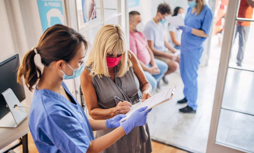 Nurse wearing a mask and gloves holding a clipboard with patient signing a document