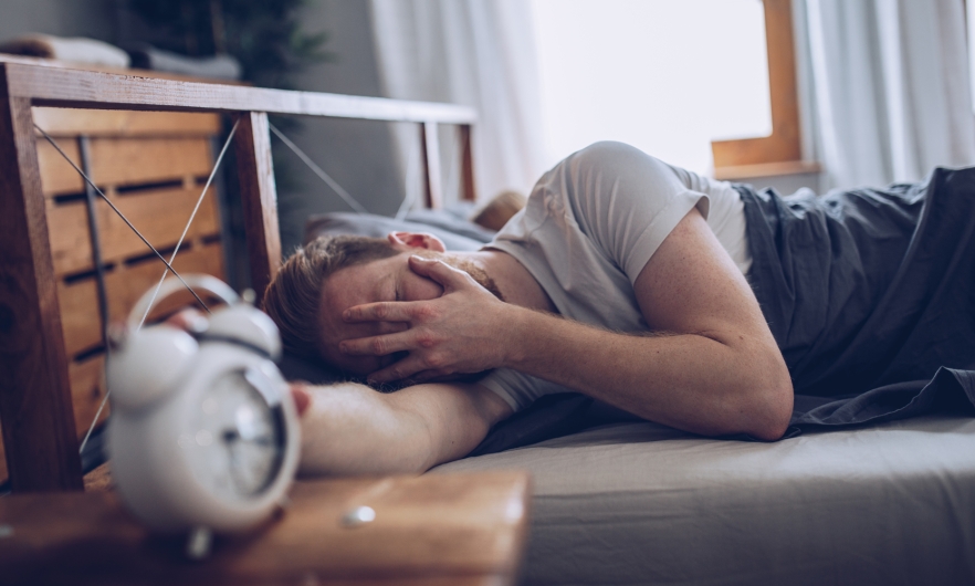 Caucasian male laying in bed covering face reaching for alarm clock