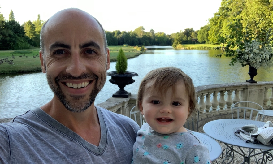 headshot of man holding young toddler with water and trees in the background