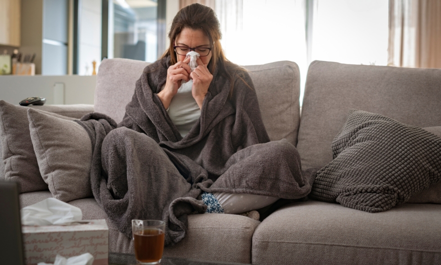 A woman sits on a couch wrapped in a blanket blowing her nose 