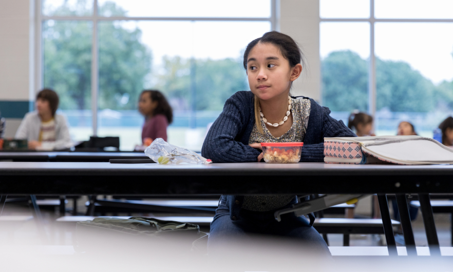 A girl sits alone in a school cafeteria with little to no food for lunch