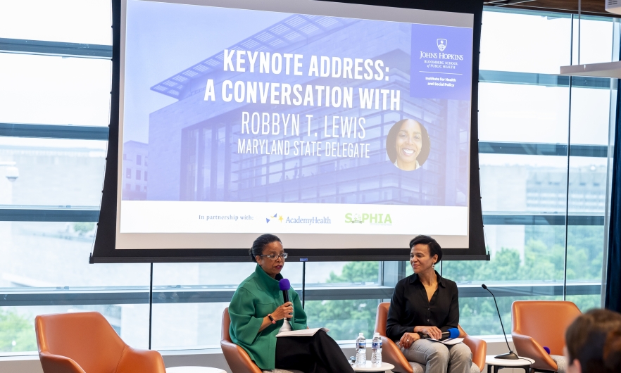 HPM Department Chair Keshia Pollack Porter and Maryland State Delegate Robbyn T. Lewis are seated in front of a projector screen that reads Keynote Address: A Conversation with Robbyn T. Lewis Maryland State Delegate