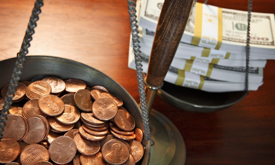 One bowl of an old fashioned weighing scale is full of pennies while the other is full of dollar bill stacks