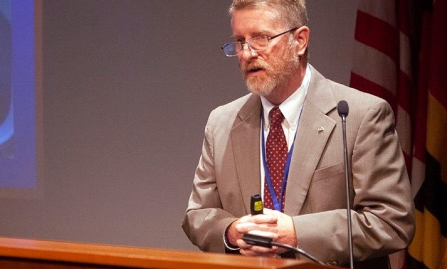 Man in tan jacket and tie standing at a podium