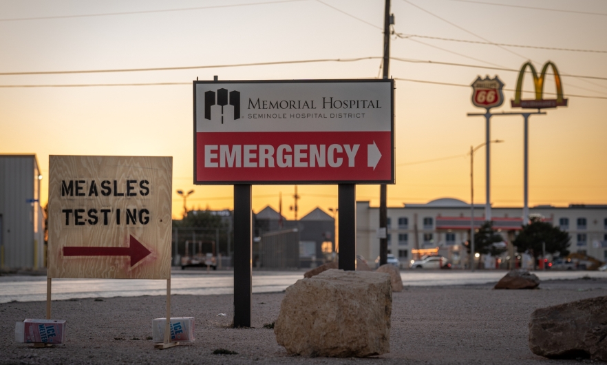 Measles testing sign in Texas A plywood sign reads "Measles Testing" and points right. Behind it, a larger permanent sign points right towards the Memorial Hospital Emergency Department.