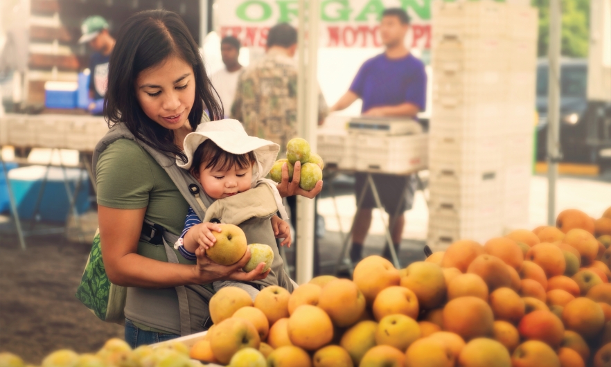 A young mom shops for fruit from organic farmers at an outdoor market. She holds her baby in front of her.