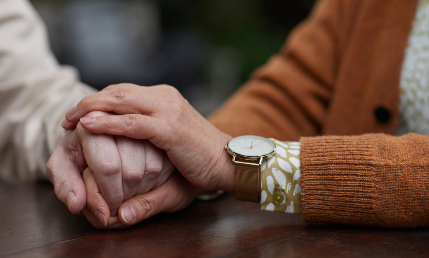 Caregiver photo A closeup of two people holding hands at a table. One of the people is younger than the other.