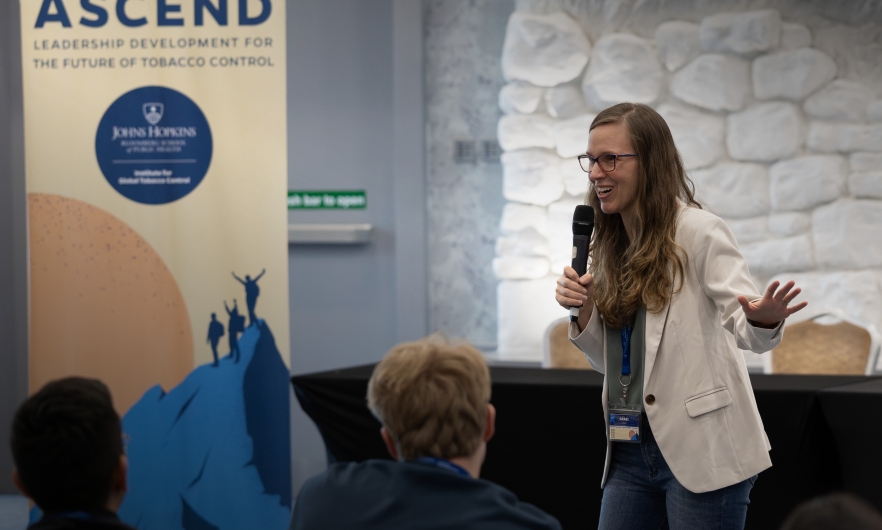 Graziele Grilo, a woman with a medium complexion and long brown hair, wearing eyeglasses and a white jacket, speaks to a room of young participants in a tobacco control leadership program