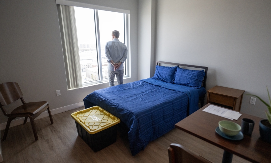 A man looks out the window of a room containing a bed, chair, small table, and storage trunk.