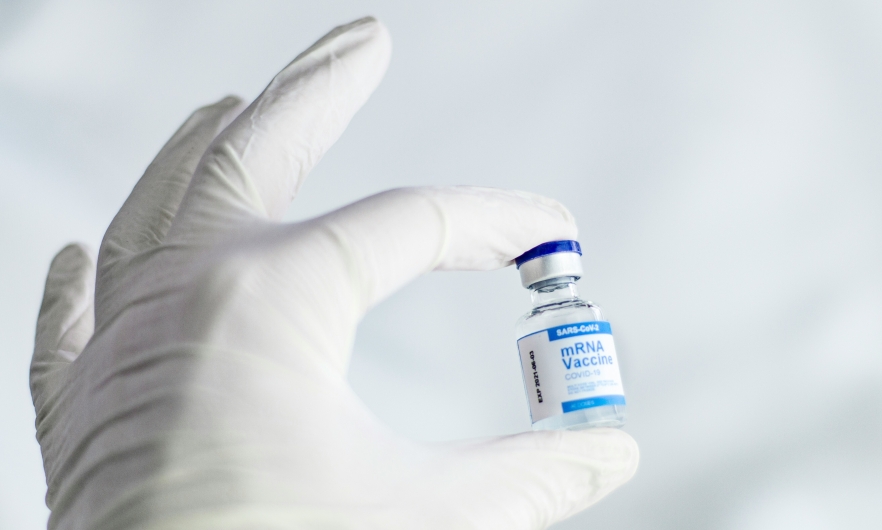 A white-gloved hand holds a vaccine vial with a blue and white label that reads "mRNA vaccine"