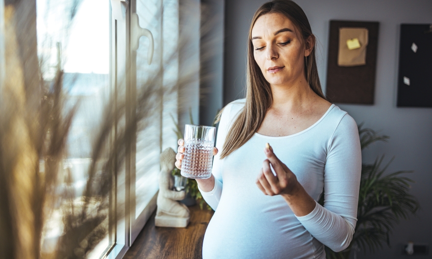 A pregnant woman with long brown hair wearing a white shirt holds a pill in one hand and a glass of water in the other. She is standing by some windows in her living room.