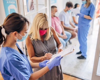 Nurse wearing a mask and gloves holding a clipboard with patient signing a document