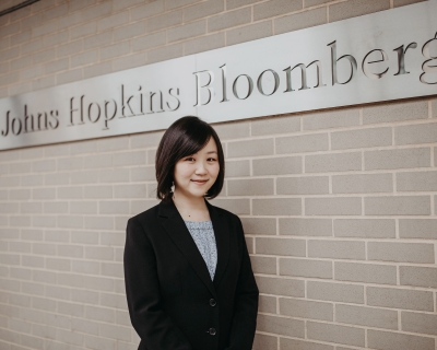 Photo of Megumi "Meg" Ichimiya standing in front of a brick wall, which features a steel signed engraved with the words "Johns Hopkins Bloomberg"