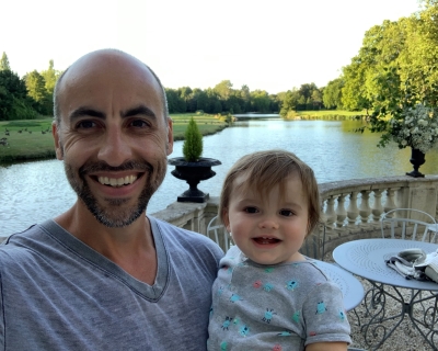 headshot of man holding young toddler with water and trees in the background