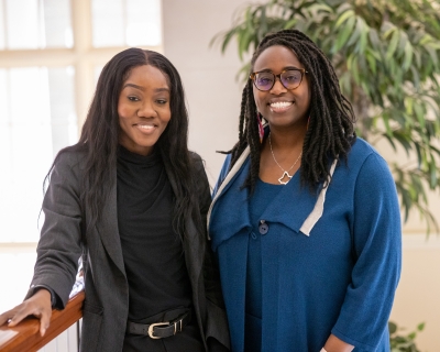 Two black women dressed in business attire smile at the camera as they stand next to each other.