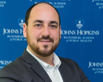 Headshot of Paul Nestadt, wearing a white collared shirt and dark gray suit jacket, in front of a blue step and repeat backdrop featuring the Bloomberg School logo
