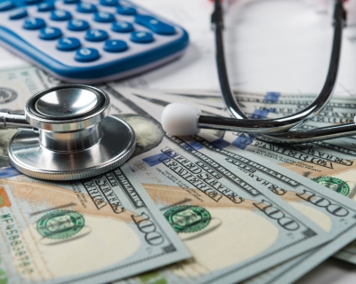 American hundred dollar bills spread out on a desk with a calculator and stethoscope placed on top.