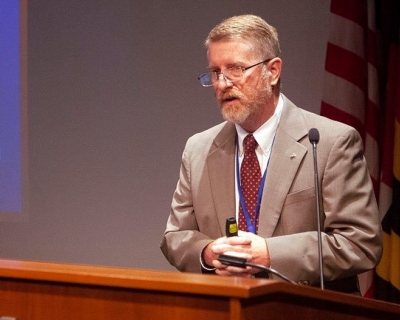 Man in tan jacket and tie standing at a podium