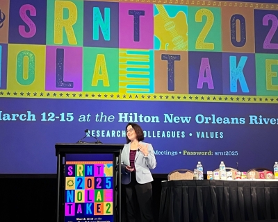 A woman in a blazer stands on stage in front of a colorful backdrop giving a presentations