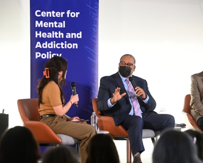 A man seated on stage speaks into his microphone.