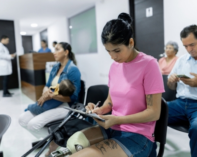 A woman with a prosthetic leg fills out forms in the waiting room of a medical office. A mother and baby, an older woman, and a middle-aged man also sit waiting.