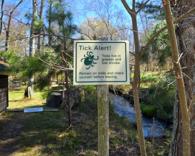 Near a wooded area of a park, a posted sign reads, Tick Alert! Ticks live in grasses and low shrubs. Remain on trails and check yourself before leaving.