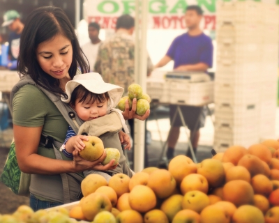 A young mom shops for fruit from organic farmers at an outdoor market. She holds her baby in front of her.