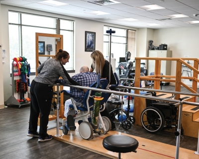 In a physical therapy office, two female therapists, one on either side, work with a man in a wheelchair who is navigating between parallel bars.