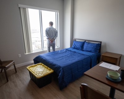 A man looks out the window of a room containing a bed, chair, small table, and storage trunk.