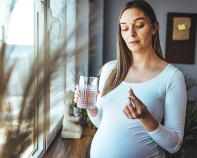 A pregnant woman with long brown hair wearing a white shirt holds a pill in one hand and a glass of water in the other. She is standing by some windows in her living room.