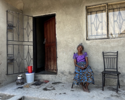 Mary Kapaipi, dressed in a bright purple and blue outfit, sits on the porch of her home with her hands clasped; a red water pitcher and bucket sits next to her door.