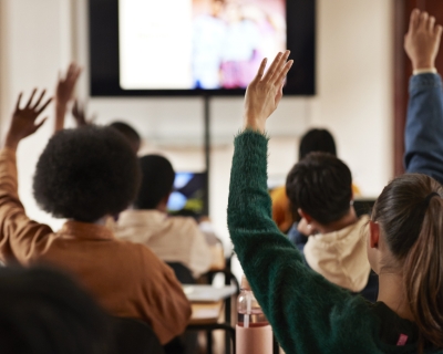 Adolescents in a classroom raising their hands, photographed from behind.
