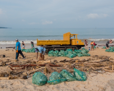 Volunteers remove plastic and other debris from a beach in Kedonganan, Indonesia; dozens of bright green trash bags holding bagged-up debris are piled up around a yellow-orange crawler dump truck.