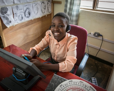 A health care worker waering a peach button-down blouse sits at a desk wearing a headset to connect with hotline callers in Lilongwe, Malawi.