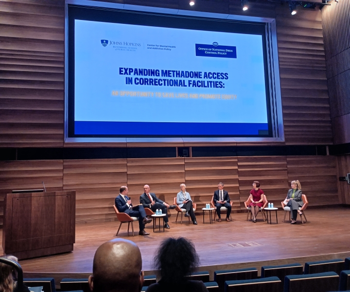 Six adults seated on an auditorium stage with a large screen visible behind them that reads Expanding Methadone Access in Correctional Facilities