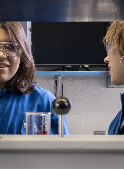 two students in blue lab coats looking at each other in a lab