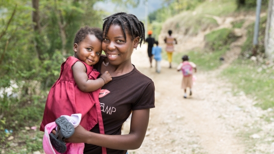 Woman holding a young child in Haiti. Both are smiling at the camera, while other children walk in the other direction down a dirt road.