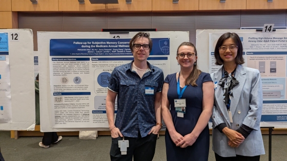 A man and two women smile as they stand in front of a research poster board. They're wearing business casual attire.