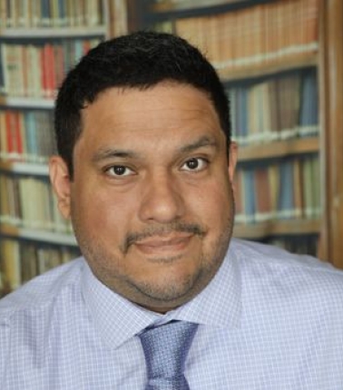 A professional headshot of a man with short dark hair and a trimmed beard, wearing a light purple checkered shirt and a light blue tie. He is standing in front of a background that appears to be a bookshelf filled with books.