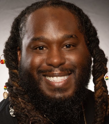 Smiling Black man with black and brown locs adorned with jewelry.