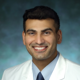 Khyzer Aziz smiles for a photo in his white coat in front of a blue photo backdrop.