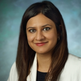 Anum Minhas smiles for a photo while wearing her white coat in front of a blue photo backdrop.