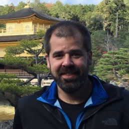 A headshot of Alden Gross, a man with a light skin tone, dark-colored mustache and beard, and very short dark hair