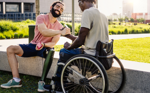 Man in wheelchair shakes hands with a smiling man with a prosthetic leg seated on stone bench outside