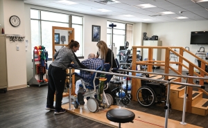 In a physical therapy office, two female therapists, one on either side, work with a man in a wheelchair who is navigating between parallel bars.